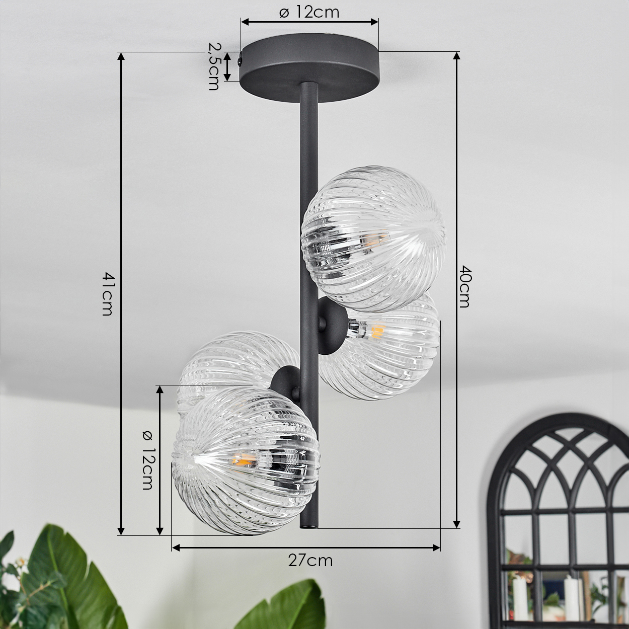 Eine moderne Deckenlampe mit drei Glaskugelschirmen. Die Lampe ist schwarz und mit Abmessungen versehen. Im Hintergrund ist ein Fenster.