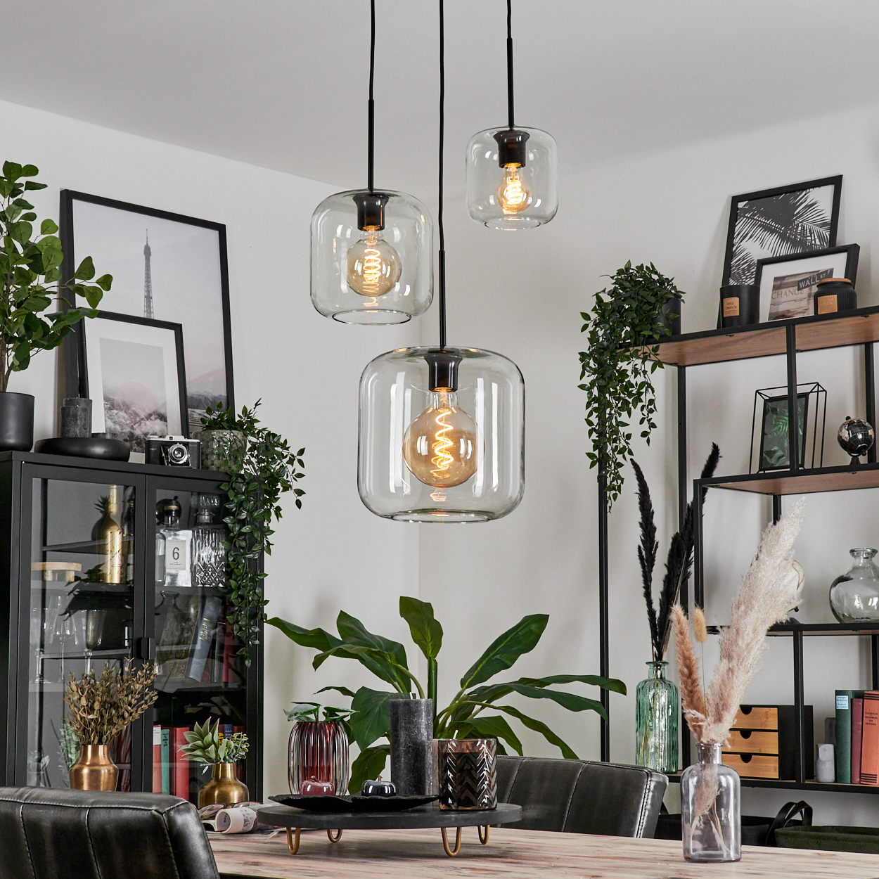 Drei Glashängeleuchten beleuchten einen Essbereich. Pflanzen, gerahmte Kunst und ein schwarzer Schrank schaffen ein stilvolles Interieur.