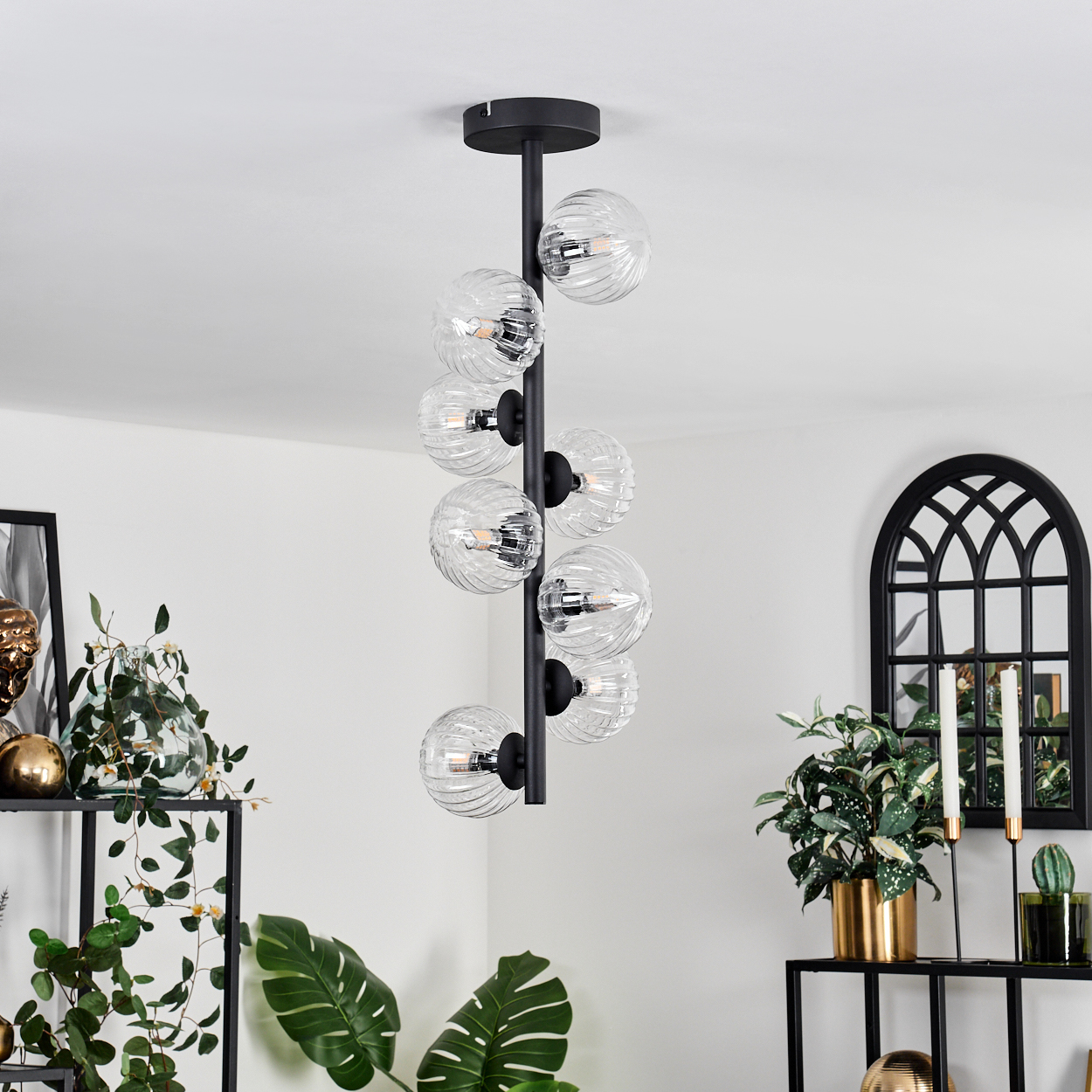 Eine moderne schwarze Pendelleuchte mit Glaskugeln hängt in einem Raum. Pflanzen und Dekorationsgegenstände sind darunter angeordnet.