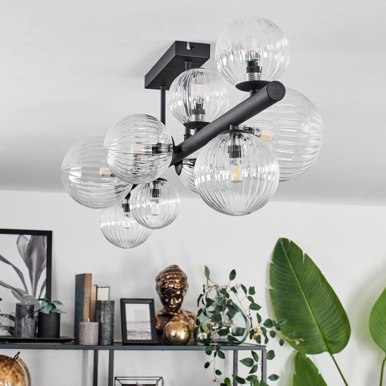 Eine moderne Deckenleuchte mit Glaskugeln hängt von der Decke. Darunter befindet sich ein Regal mit Dekorationen, darunter eine Büste und Pflanzen.