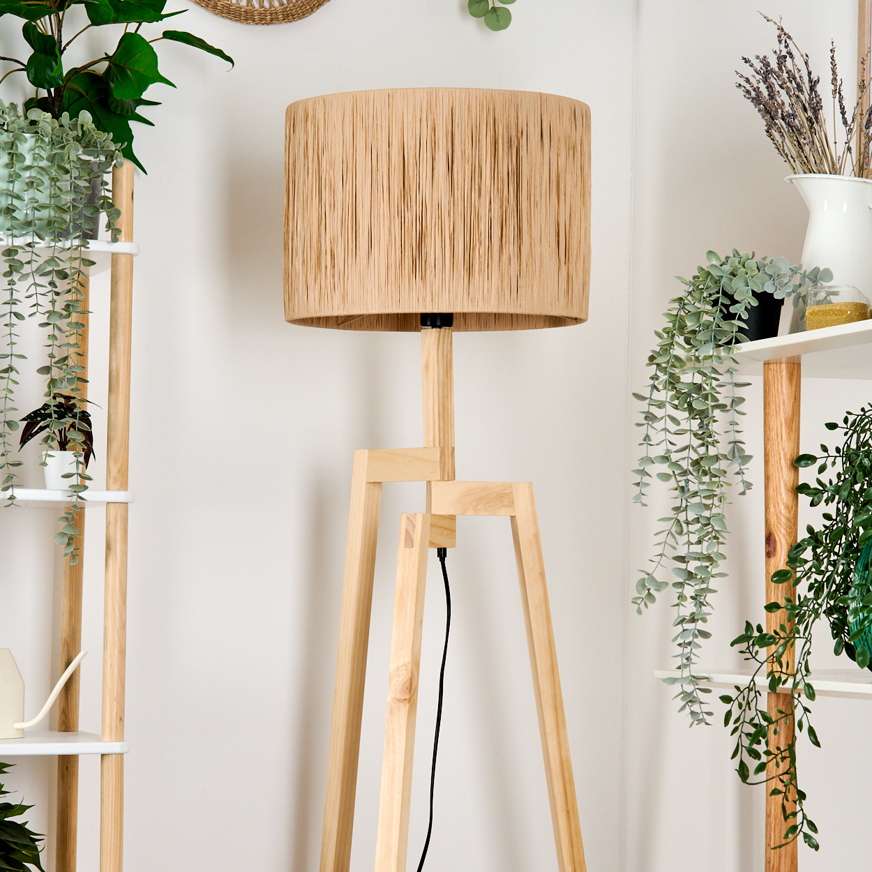 Beigefarbener Lampenschirm auf einem Holzstativ, umgeben von Pflanzen auf Regalen und einer weißen Wand im Hintergrund.