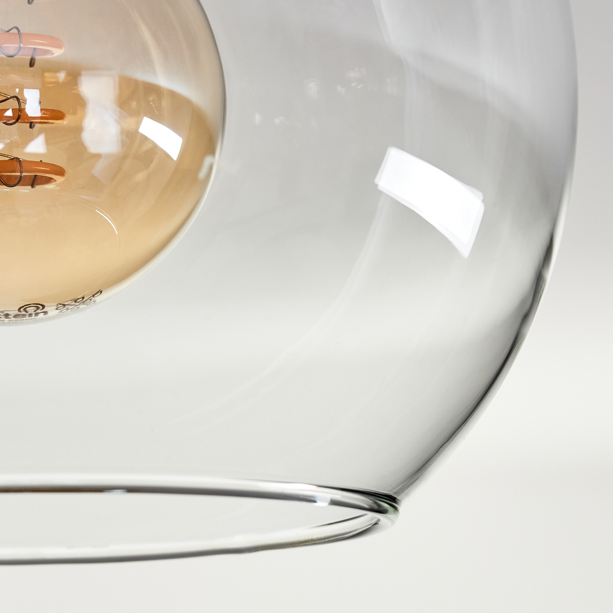 Nahaufnahme eines Lampenschirms aus klarem Glas mit einer warm getönten Glühbirne. Der Hintergrund ist eine weiße Oberfläche.
