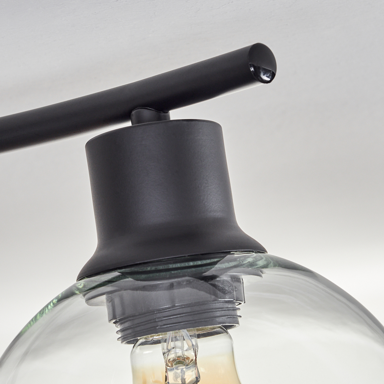 Nahaufnahme einer schwarzen Lampenhalterung mit Glasschirm und sichtbarer Glühbirne. Der Hintergrund ist eine weiße Decke.