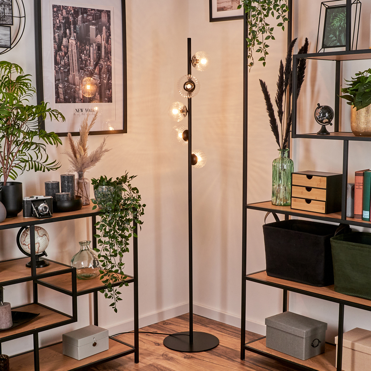 Eine Zimmerecke mit Regalen, einer Stehlampe und Dekoration. Regale halten Pflanzen, Bücher und Kisten. Die Lampe hat Glaskugellampen.