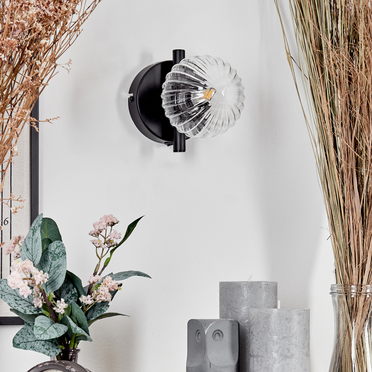 Eine dekorative Wandleuchte mit Glasschirm ist an einer weißen Wand montiert. Getrocknete Pflanzen und Blumen sind in Vasen. Graue Kerzen und Deko.