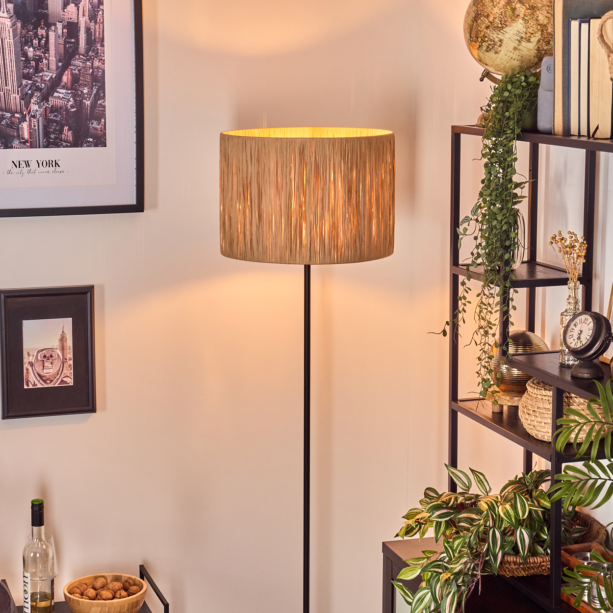 Eine beleuchtete Stehlampe mit beigefarbenem Lampenschirm steht neben einem Bücherregal mit Pflanzen und Dekorationsartikeln. Gemälde hängen an der Wand.