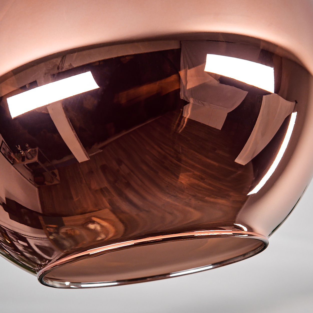 Eine kupferfarbene, reflektierende Leuchte. Sie reflektiert einen Raum mit Holzböden und weißen Wänden. Helle rechteckige Lichter werden reflektiert.