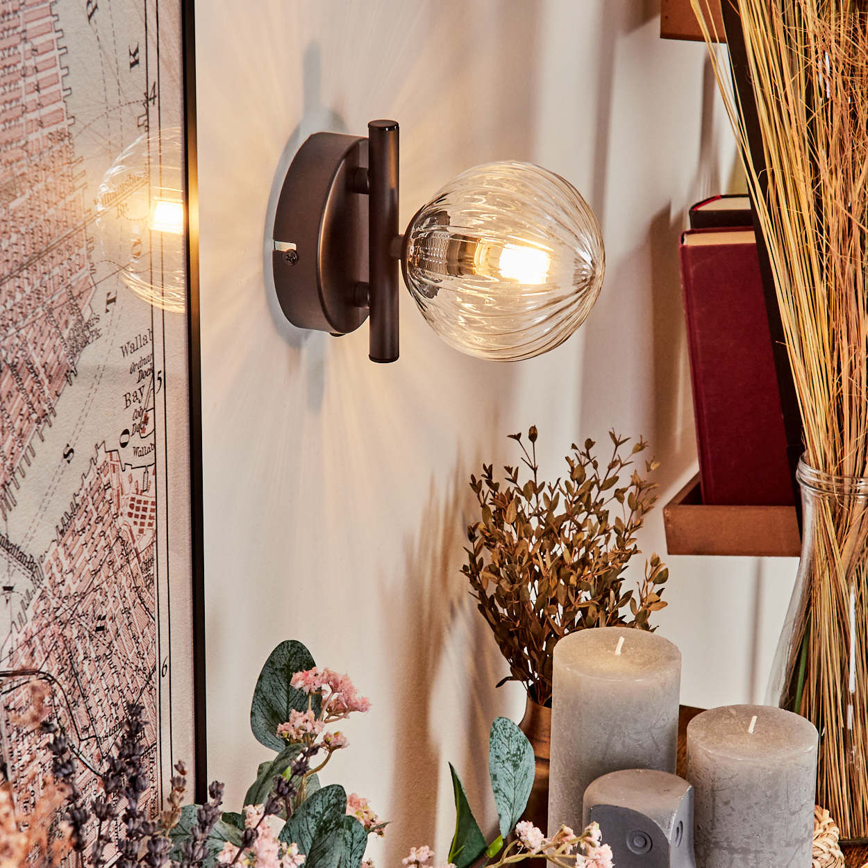 Eine dekorative Wandlampe mit Glaskugel. Kerzen und Blumen sind vor einer Karte und Büchern platziert.