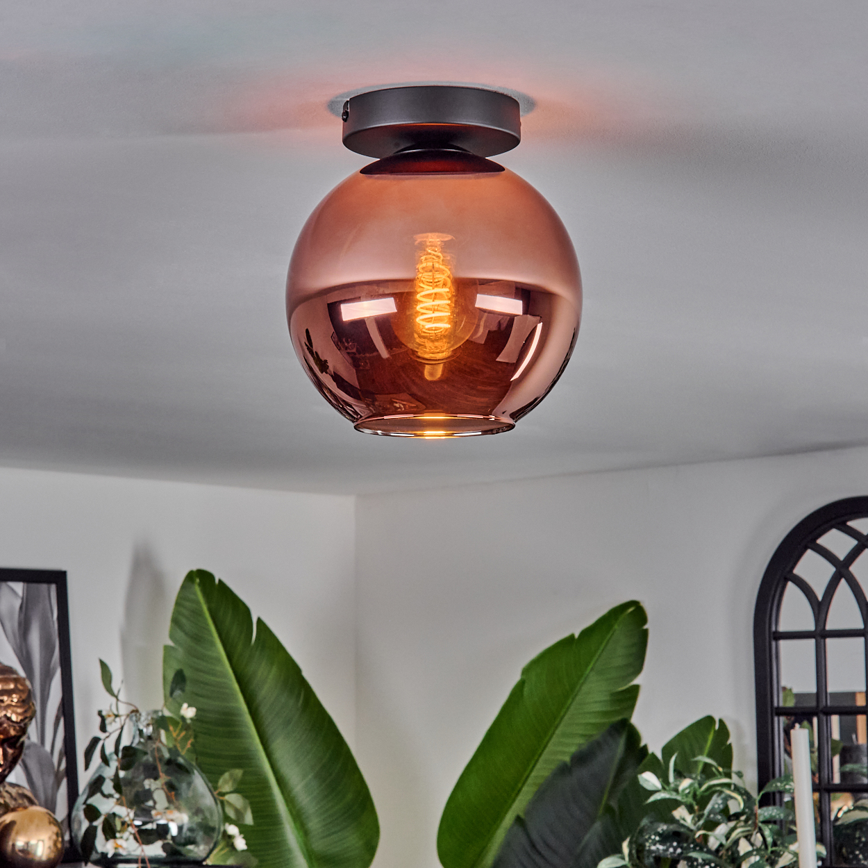 Kupferfarbene Deckenlampe aus Glas mit sichtbarer Glühbirne. Grüne Pflanzen im Vordergrund. Weiße Wand im Hintergrund.