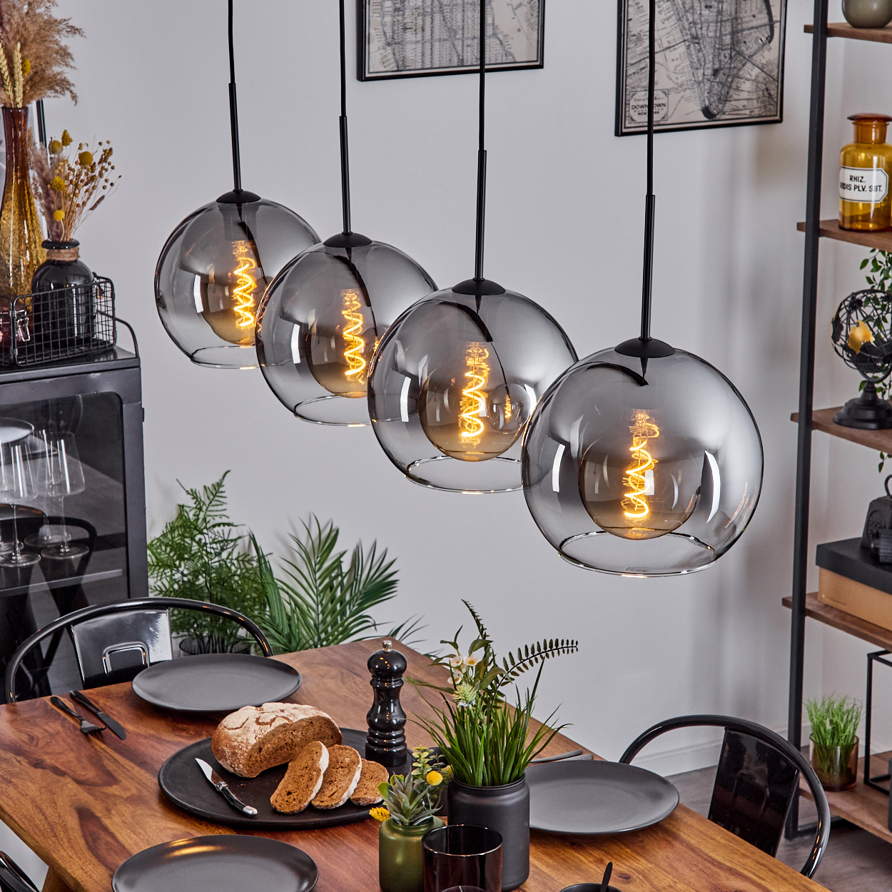 Esstisch mit Tellern, Brot und Pflanzen. Vier Kugelpendelleuchten hängen darüber. Regale mit Gegenständen im Hintergrund.