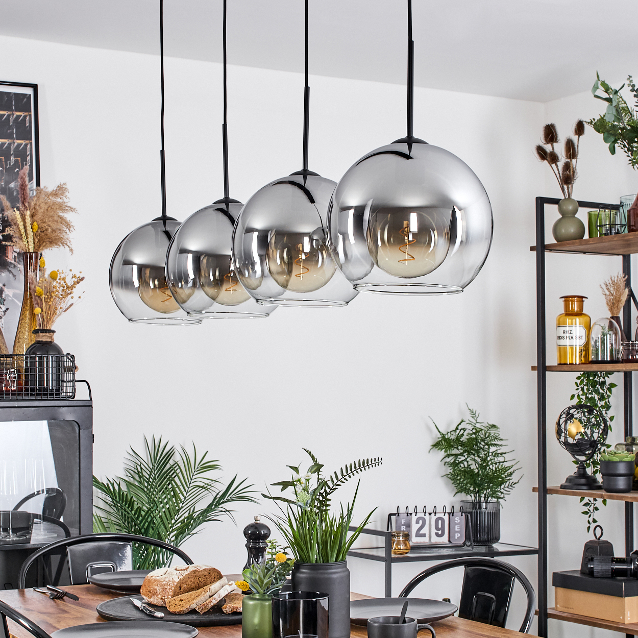 Esszimmer mit Hängelampen, Tisch mit Brot, Pflanzen und Kalender. Regale mit Dekoration im Hintergrund.
