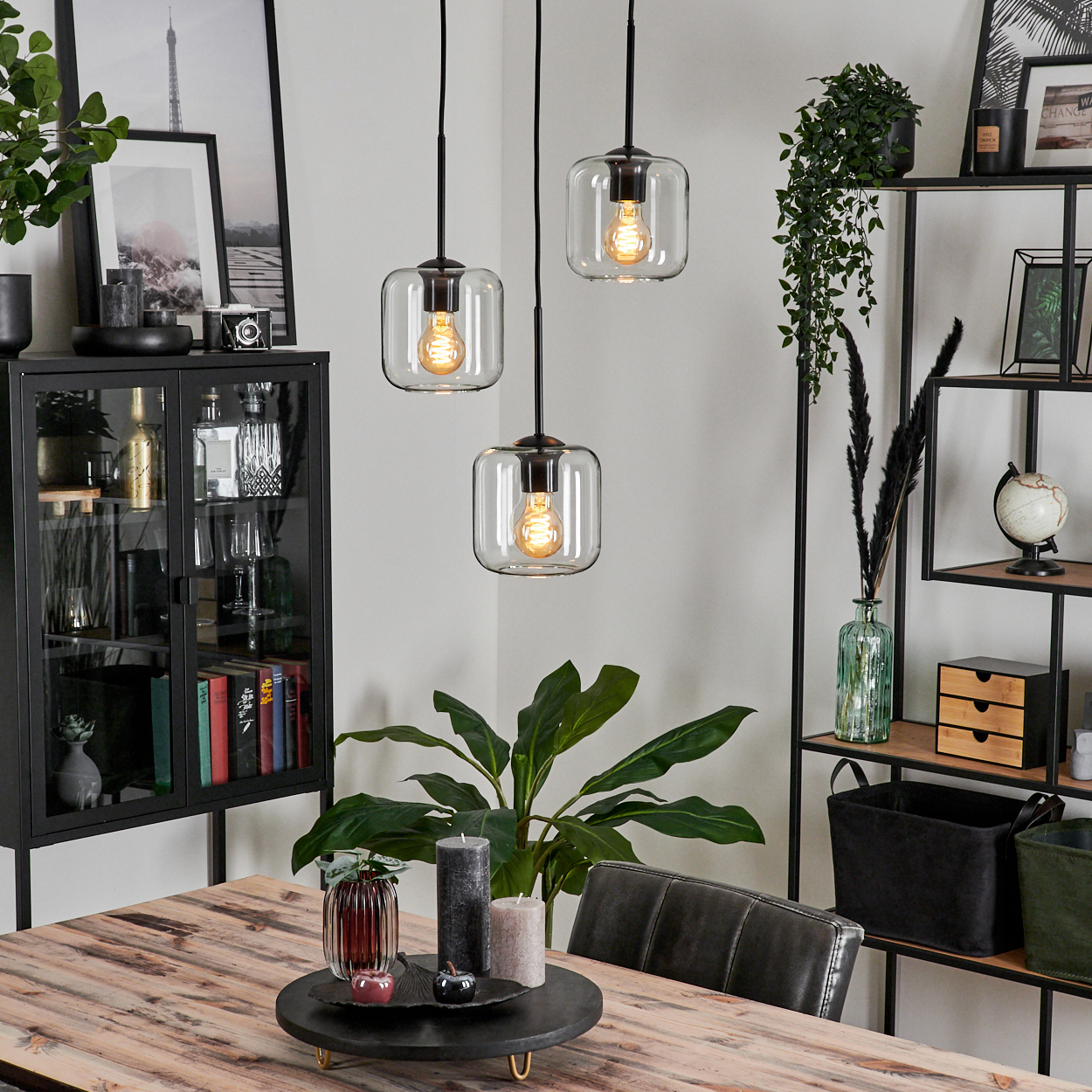 Drei Pendelleuchten hängen über einem Holztisch, der mit Kerzen und einer Vase dekoriert ist. Ein schwarzer Schrank und Regal sind im Hintergrund.