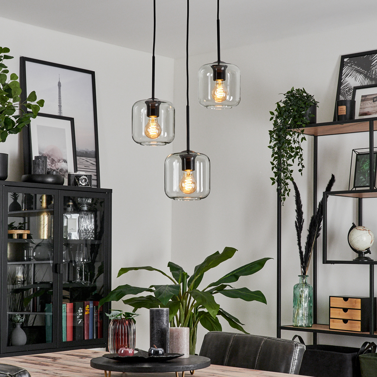 Esszimmer mit drei Pendelleuchten. Enthält Pflanzen, einen Schrank und einen Holztisch. Der Hintergrund hat Regale und ein gerahmtes Foto.