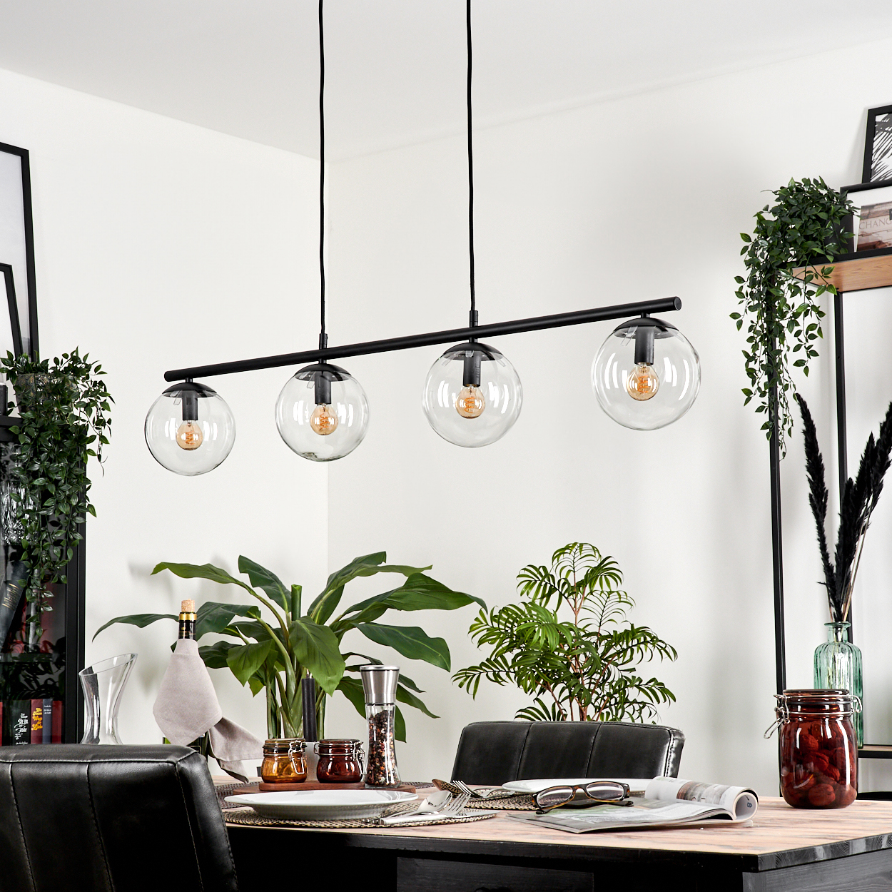 Ein Esszimmer mit einem gedeckten Tisch. Eine schwarze Hängelampe hängt darüber. Pflanzen und Dekoration sind sichtbar.