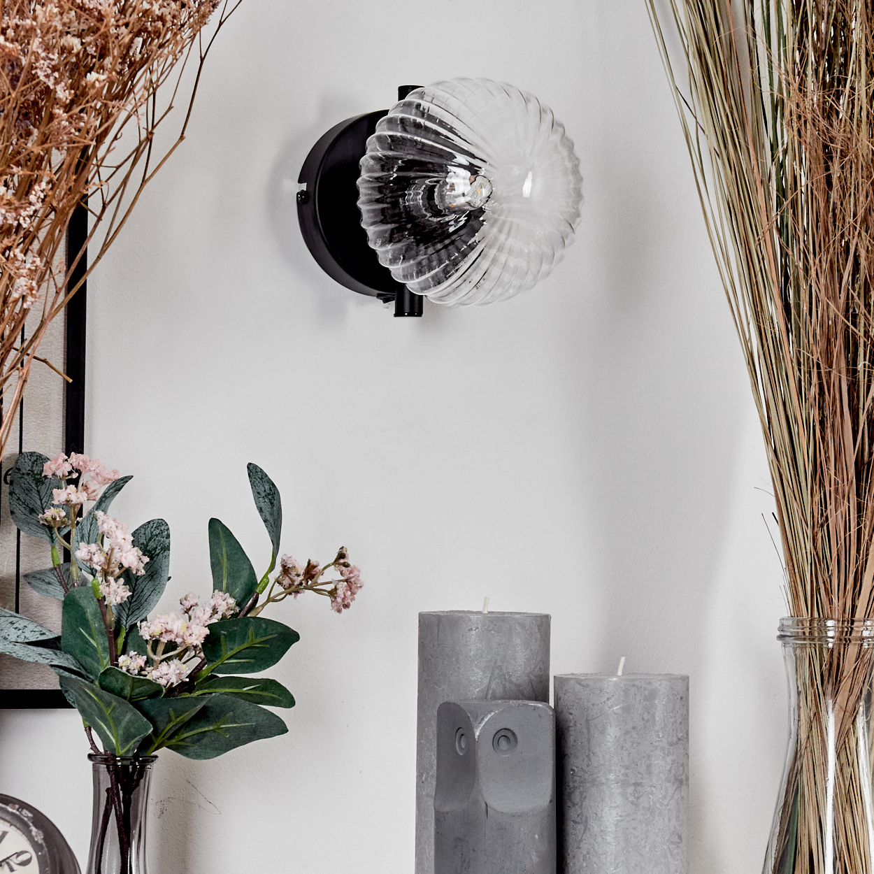 Eine dekorative Wandleuchte mit Glasschirm. Es gibt Kerzen, eine Vase mit trockenem Gras und Blumen.