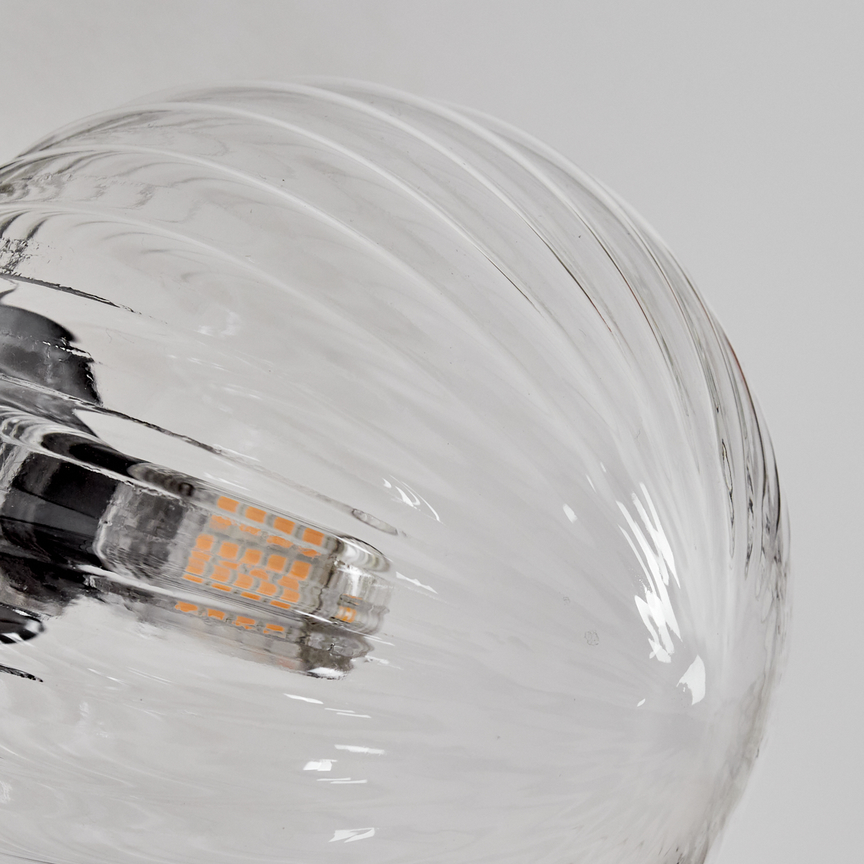 Nahaufnahme einer durchsichtigen Glasglühbirne. Die Glühbirne hat ein geripptes Design. Im Inneren befindet sich eine kleine orangefarbene Lichtquelle. Der Hintergrund ist weiß.