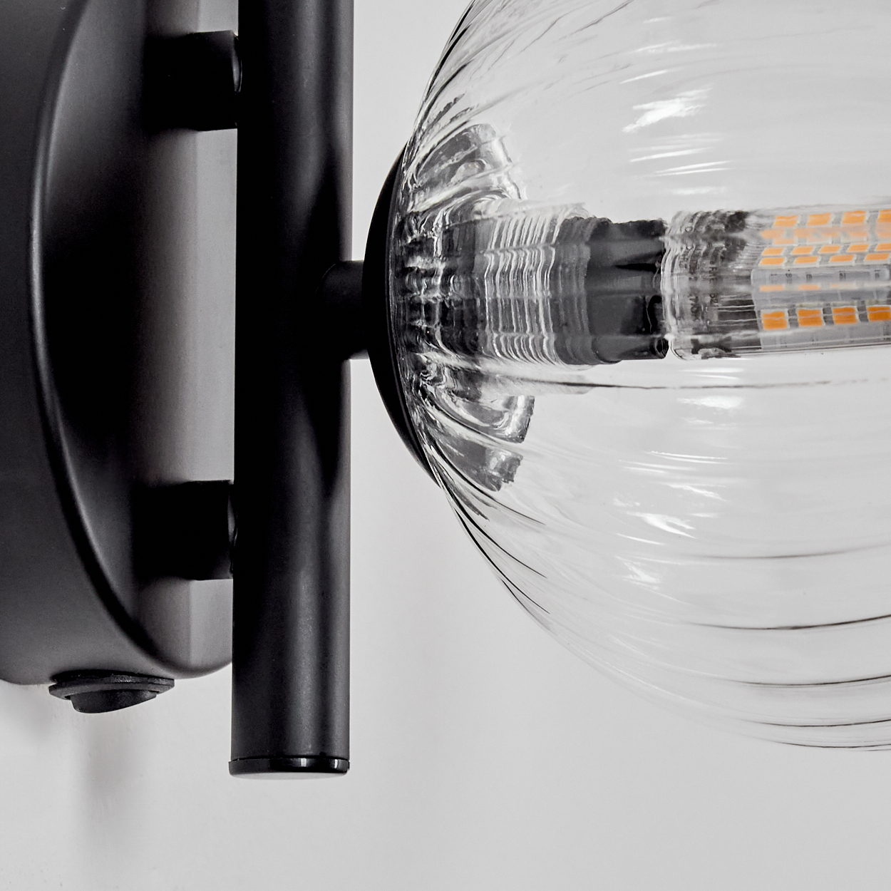 Nahaufnahme einer Wandleuchte. Ein schwarzer Metallrahmen trägt eine gerippte Glaskugel mit LED-Licht. Der Hintergrund ist weiß.