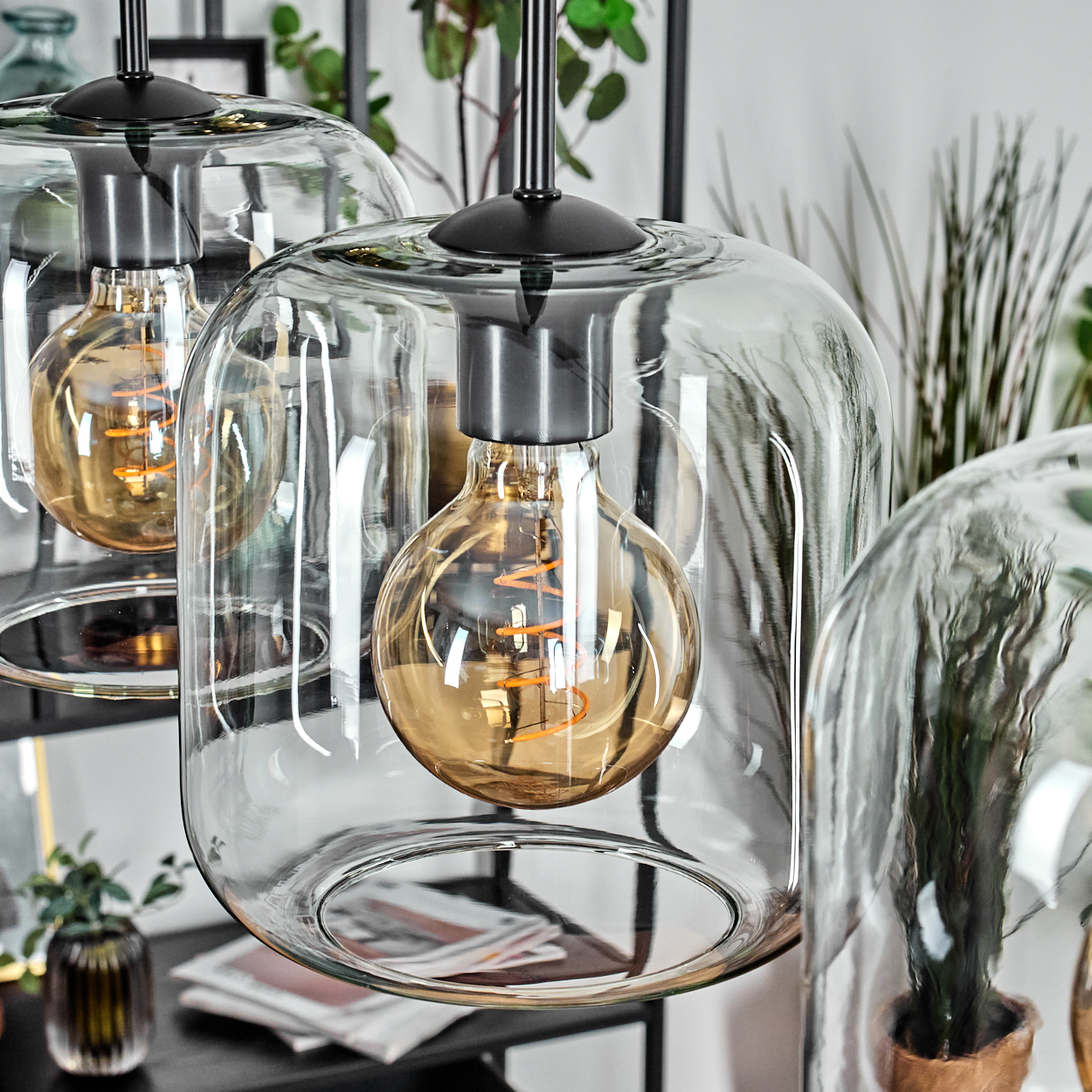 Drei Glas-Pendelleuchten mit Edison-Glühbirnen hängen. Pflanzen und Dekoration sind im Hintergrund.