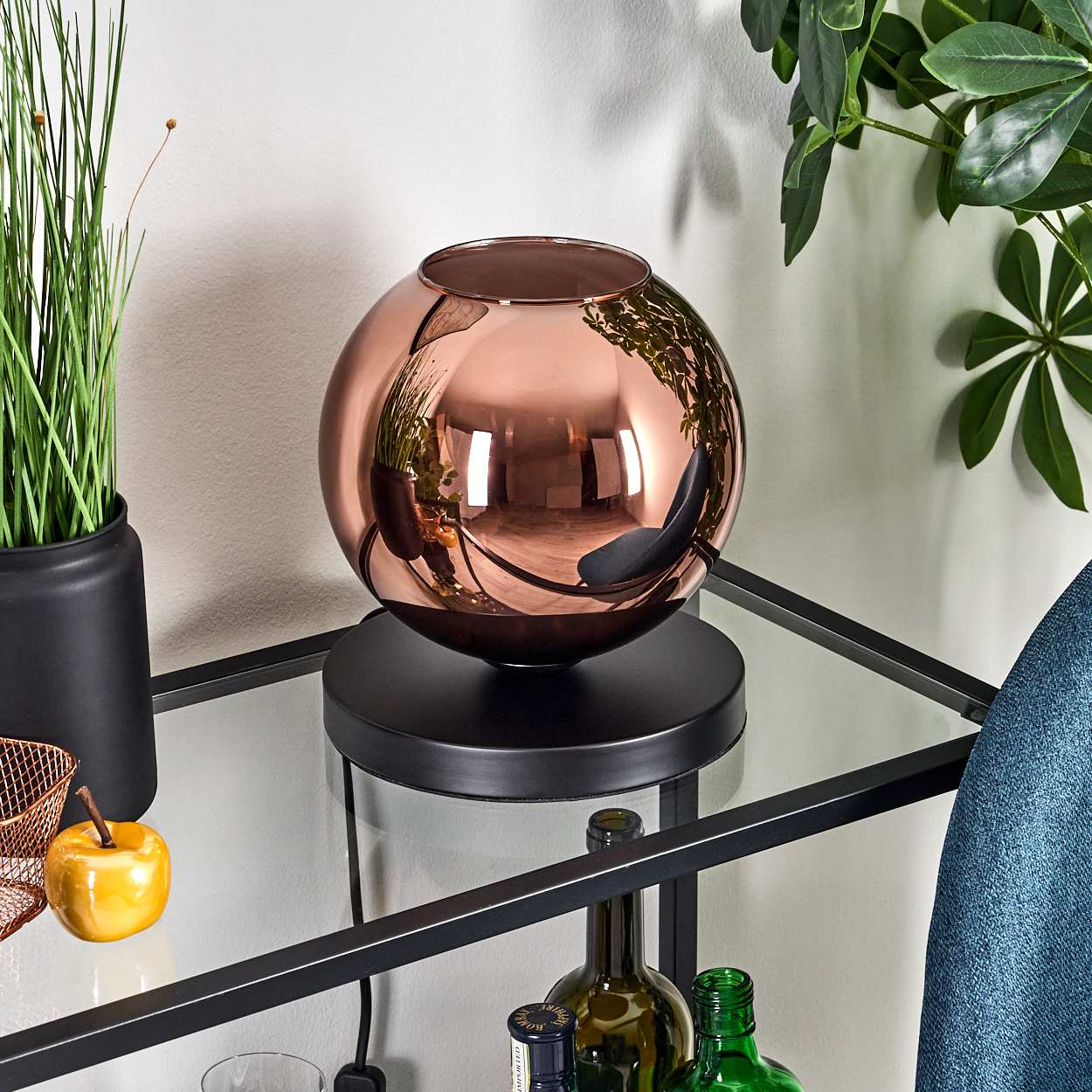 Eine kupferfarbene Kugellampe auf einem schwarzen Ständer reflektiert Objekte. Pflanzen und ein Apfel stehen auf dem Regal. Im Hintergrund ist eine weiße Wand zu sehen.