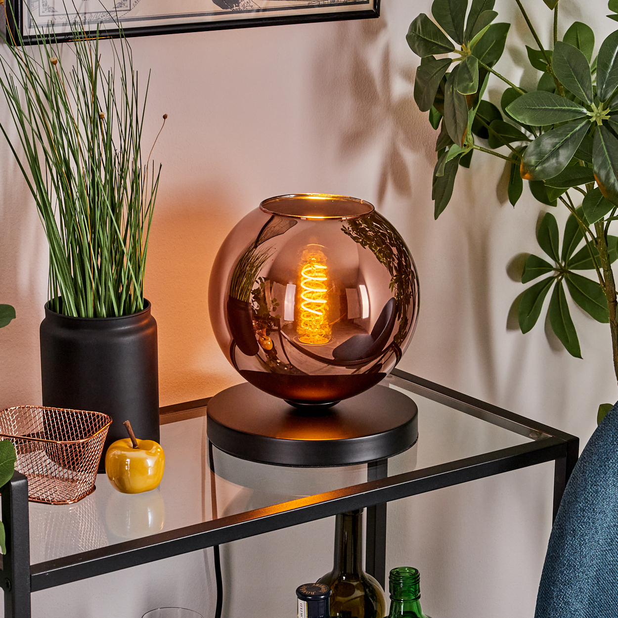Eine dekorative Lampe mit kupferfarbenem Glasschirm, beleuchteter Glühbirne und schwarzem Sockel auf einem Glastisch. Pflanzen und Dekor umgeben es.