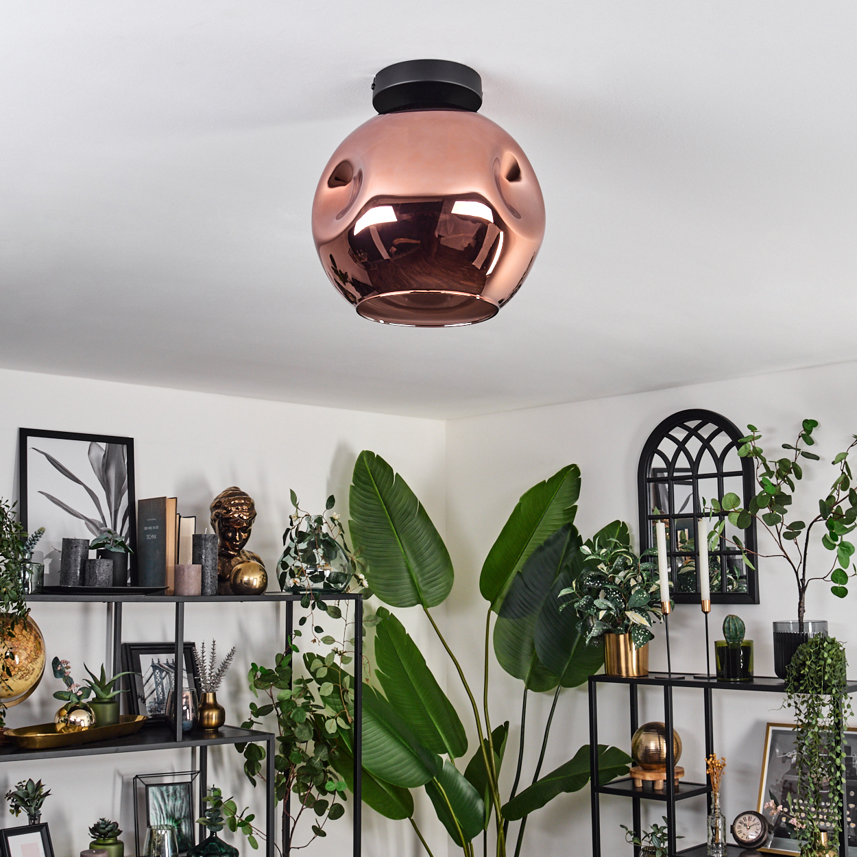 Eine kupferfarbene Deckenlampe hängt über einem Regal mit Pflanzen, Büchern und Dekoration. Große grüne Blätter.