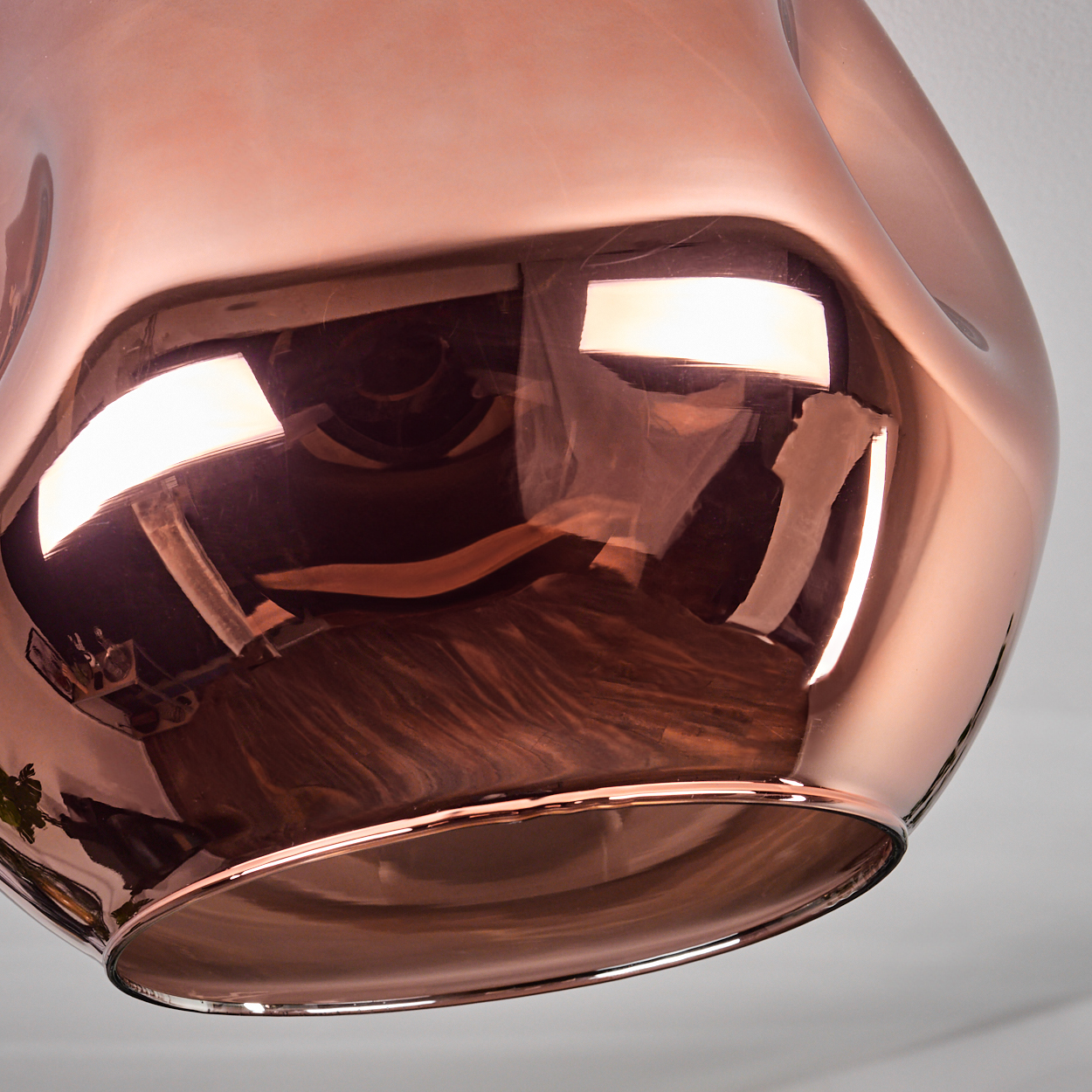 Eine kupferfarbene, reflektierende Pendelleuchte hängt von einer weißen Decke. Die Oberfläche der Leuchte reflektiert die Umgebung.