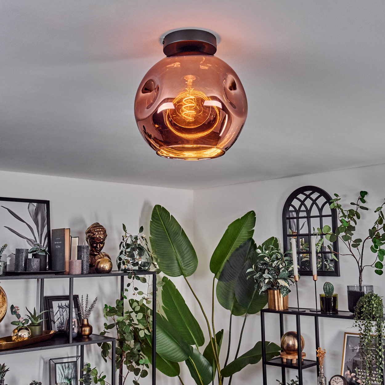 Dekorative Deckenlampe mit kupferfarbenem Glasschirm. Darunter Regale mit Pflanzen, Kerzen und Dekoration, die eine gemütliche Atmosphäre schaffen.