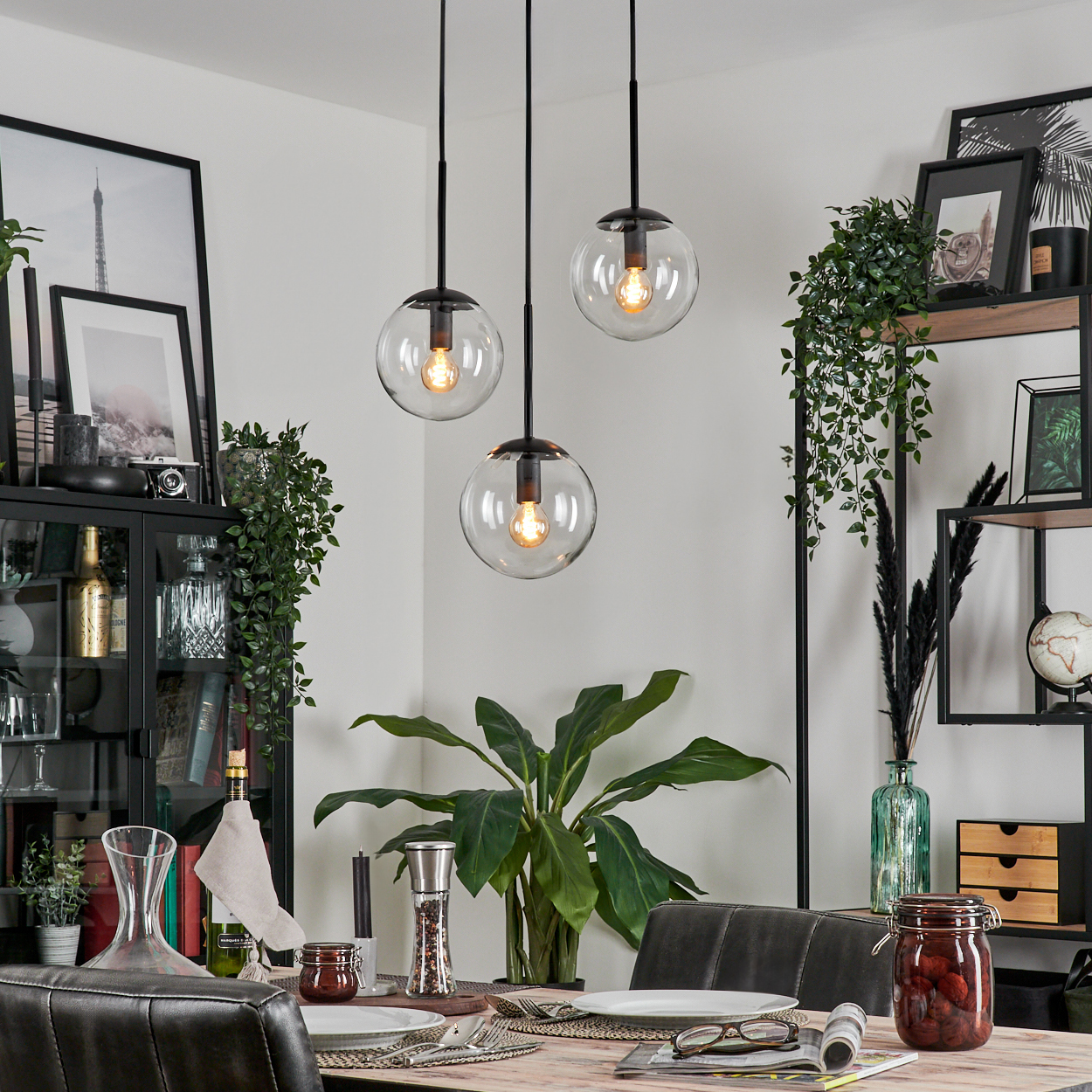 Ein Esszimmer mit einem gedeckten Tisch. Drei Pendelleuchten hängen darüber. Pflanzen und Dekoration sind im Raum angeordnet.
