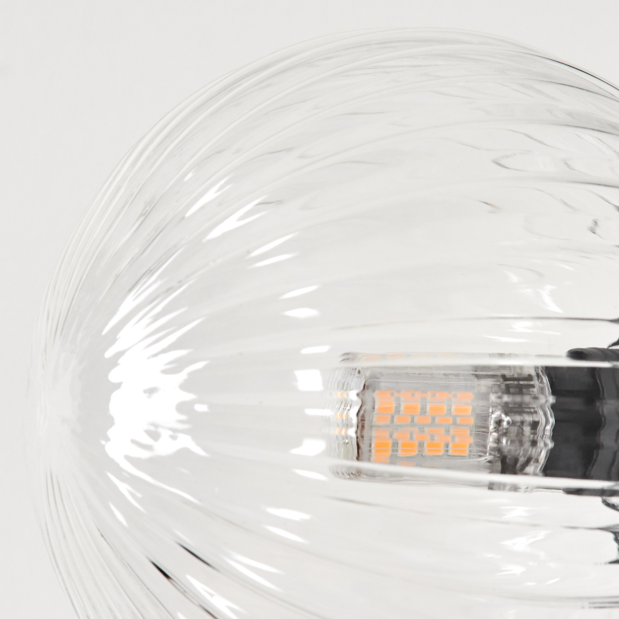 Nahaufnahme eines Lampenschirms aus klarem Glas mit einer Glühbirne. Die Glühbirne hat orangefarbene Lichter. Weißer Hintergrund. Glas hat strukturierte Linien.