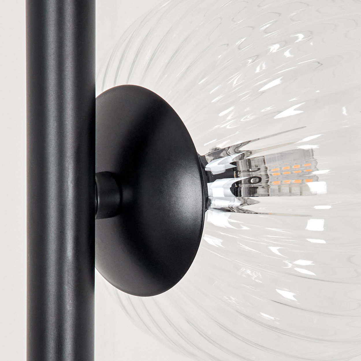 Nahaufnahme einer schwarzen Lampenhalterung. Eine transparente, strukturierte Glühbirne ist befestigt. Eine schwarze Stange stützt die Halterung vor weißem Hintergrund.