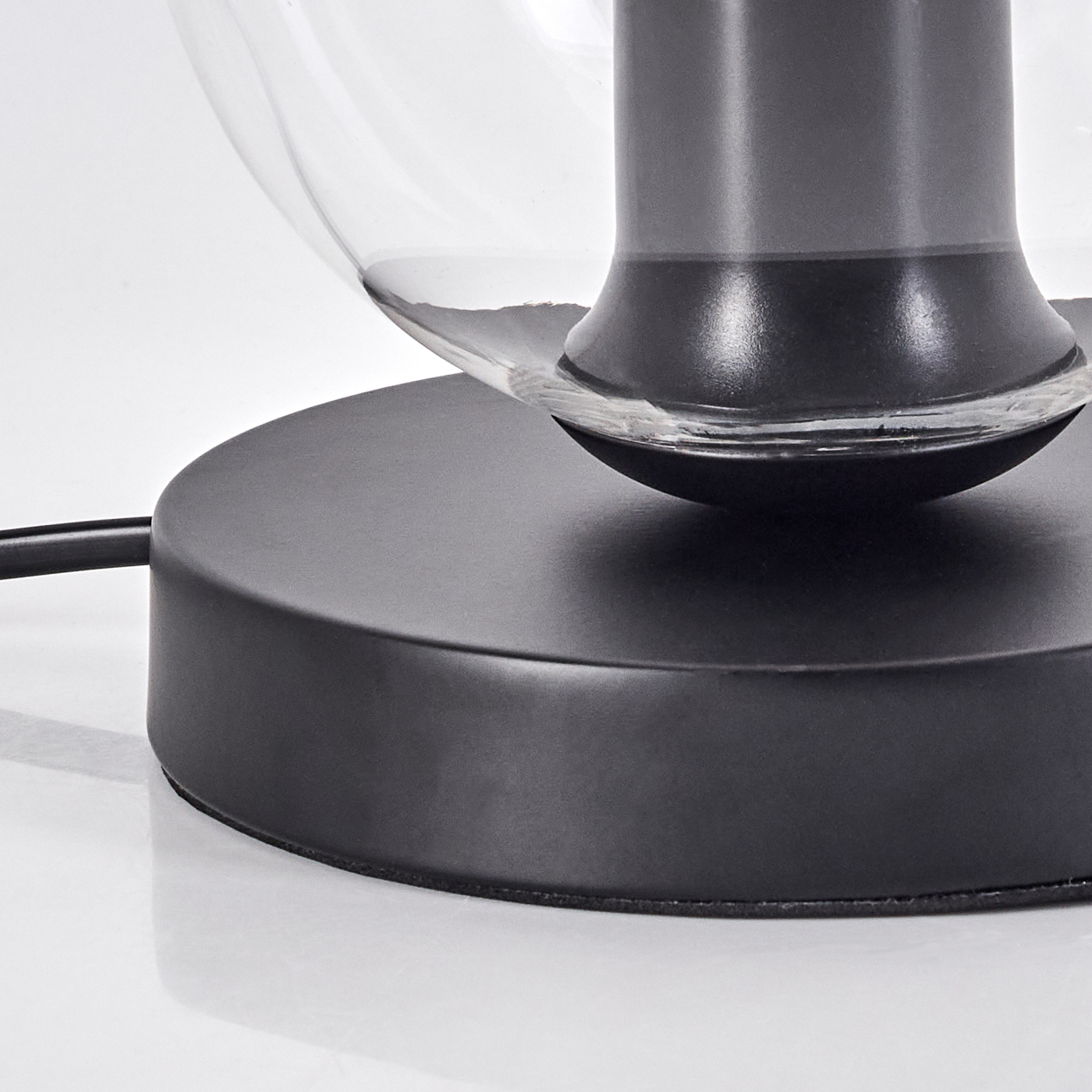 Nahaufnahme eines schwarzen Lampenfußes mit einem transparenten Glasschirm. Ein schwarzes Kabel erstreckt sich vom Sockel. Der Hintergrund ist weiß.