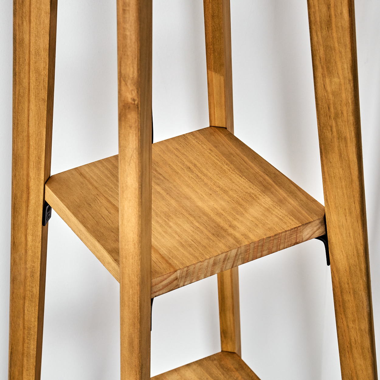 Pflanzenständer aus Holz mit drei Regalen. Hellbraunes Holz, schlichtes Design. Weißer Wandhintergrund. Schwarze Metallstützen.