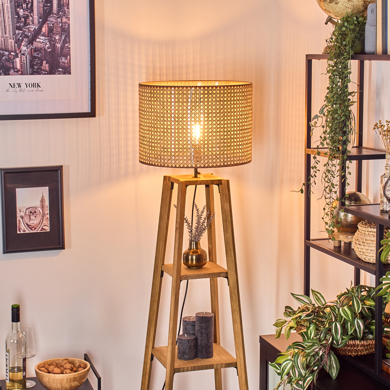 Eine Holzlampe mit Wicker-Schirm steht in einem Raum. Regale mit Pflanzen und Dekoration befinden sich im Hintergrund. Kunstwerke und Kerzen sind ebenfalls vorhanden.