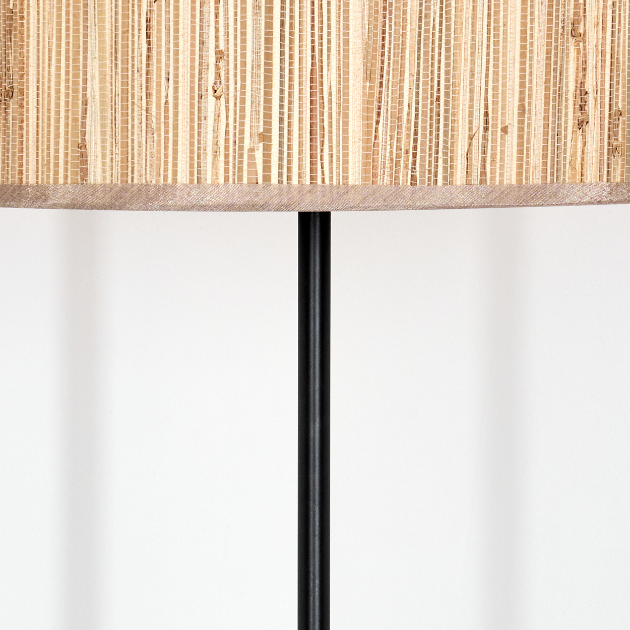 Eine Lampe mit gewebtem Lampenschirm und schwarzem Metallpfosten. Der Hintergrund ist eine weiße Wand mit Schatten.