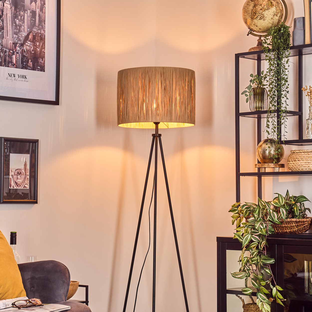 Eine Stehlampe steht in einem Raum mit Dekoration. Ein Regal mit Pflanzen und einem Globus, gerahmte Bilder an der Wand. Ein Sofa mit gelbem Kissen.