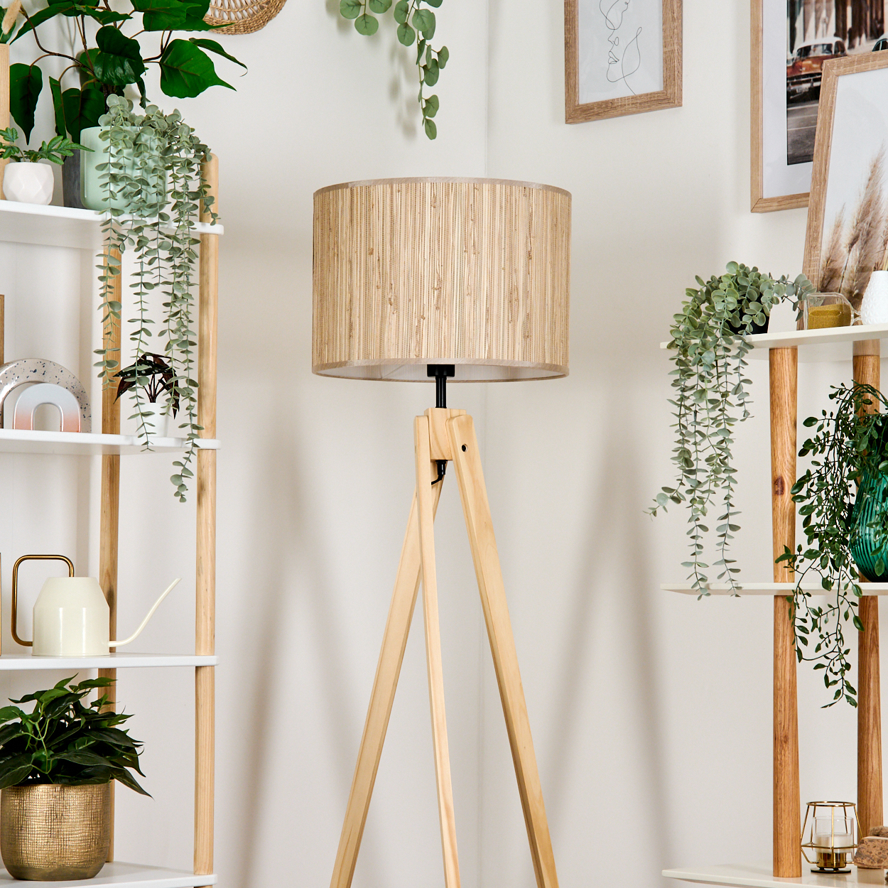 Eine Holz-Stativlampe mit beigem Lampenschirm steht in einer Ecke mit Regalen, die Pflanzen und gerahmte Bilder an einer weißen Wand tragen.