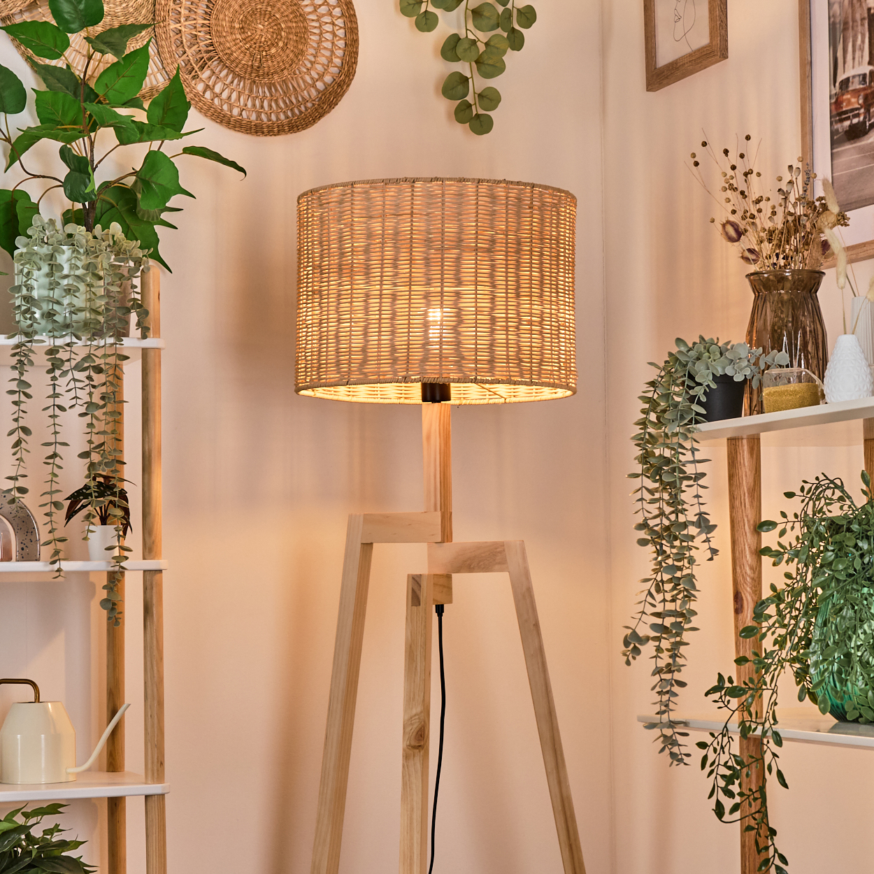 Stehlampe aus Holz mit gewebtem Lampenschirm, umgeben von Pflanzen auf Regalen und von der Wand hängend. Warme, weiche Beleuchtung.