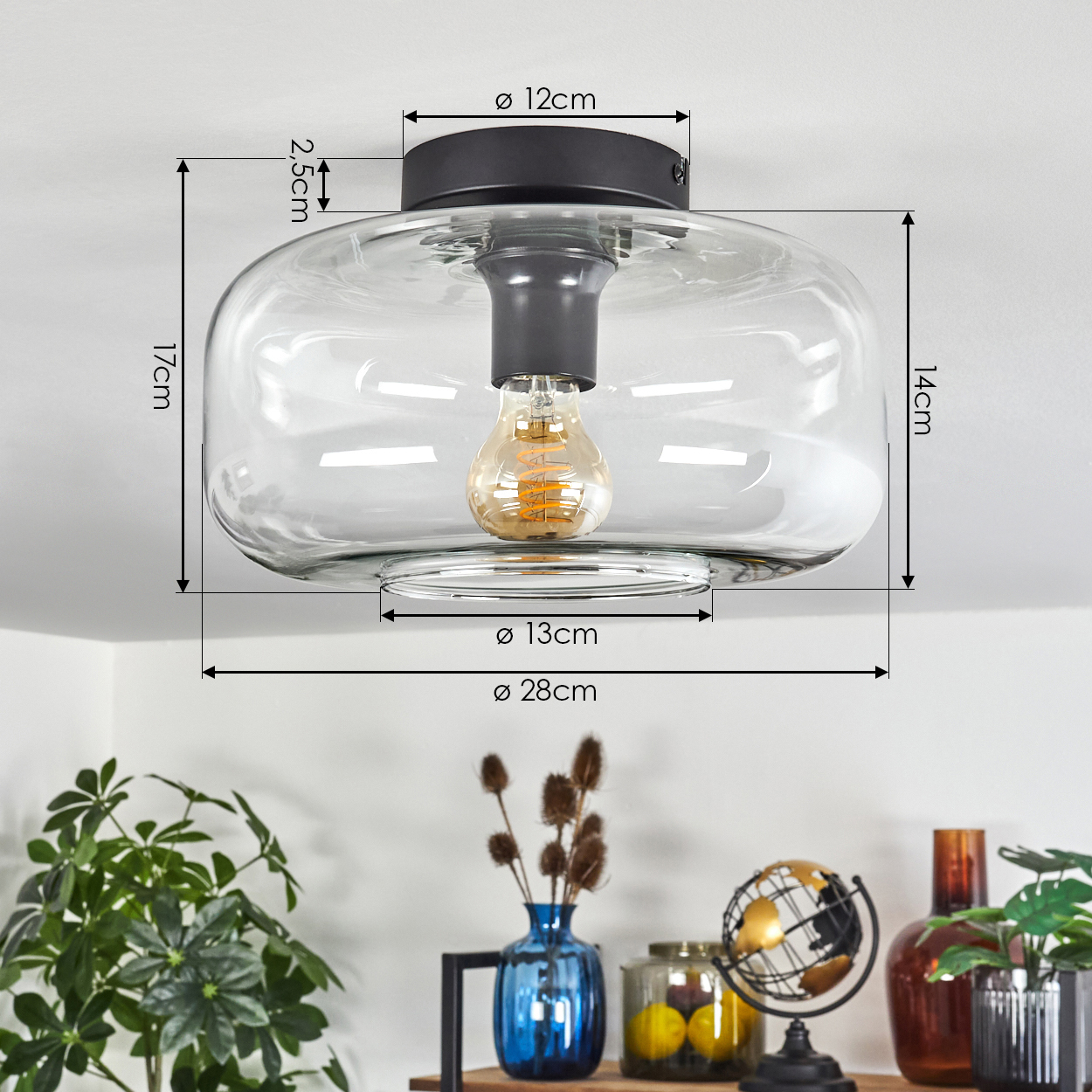Eine transparente Glasdeckenlampe mit schwarzem Sockel und sichtbarer Glühbirne, mit Maßangaben. Darunter ein Regal mit Deko.