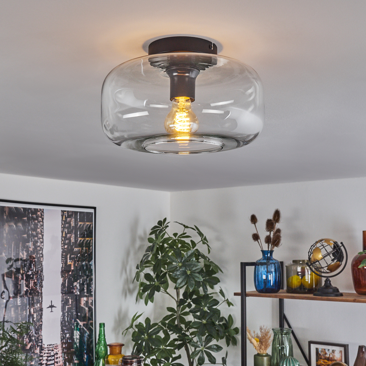 Eine moderne Deckenleuchte mit Glasschirm und sichtbarer Glühbirne. Im Hintergrund sind Pflanzen und Dekoration zu sehen.