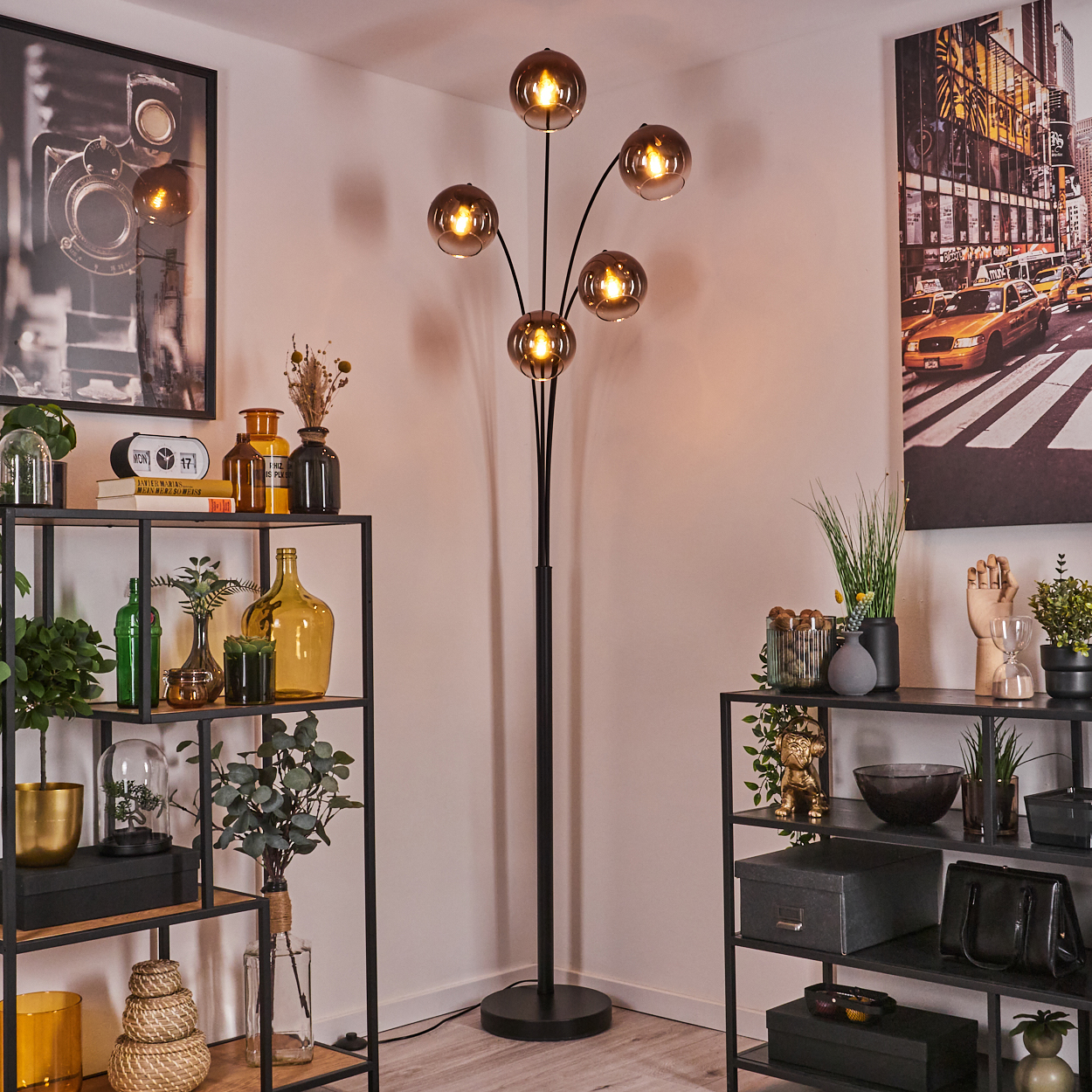 Eine Zimmerecke mit einer schwarzen Stehlampe mit Glaskugeln, Regalen mit Dekor und Bildern an der Wand.