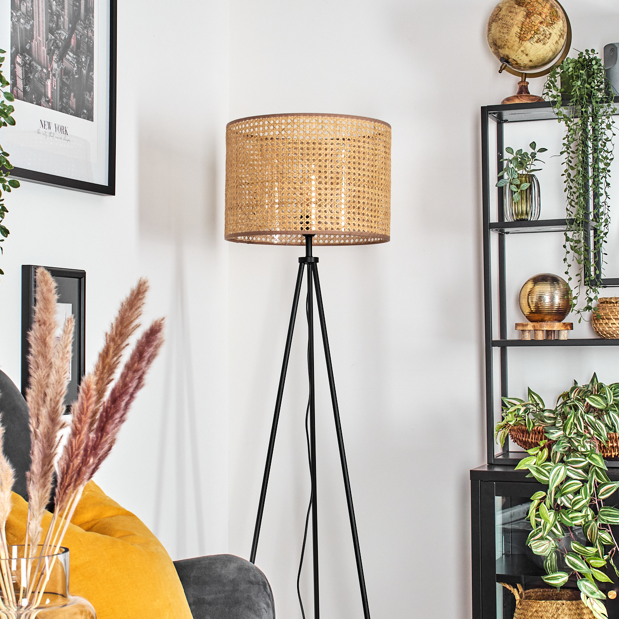 Eine gewebte Lampe auf einem schwarzen Stativ, ein schwarzes Regal mit Pflanzen und Globus, ein New York Bild und Pampasgras.