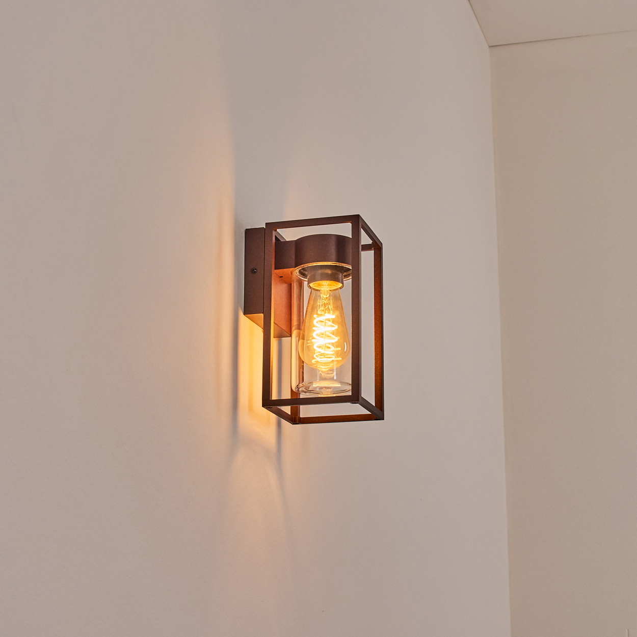 Eine Wandleuchte mit braunem Rahmen und einer Glühbirne mit Spiralfaden, die warmes Licht auf eine weiße Wand wirft.