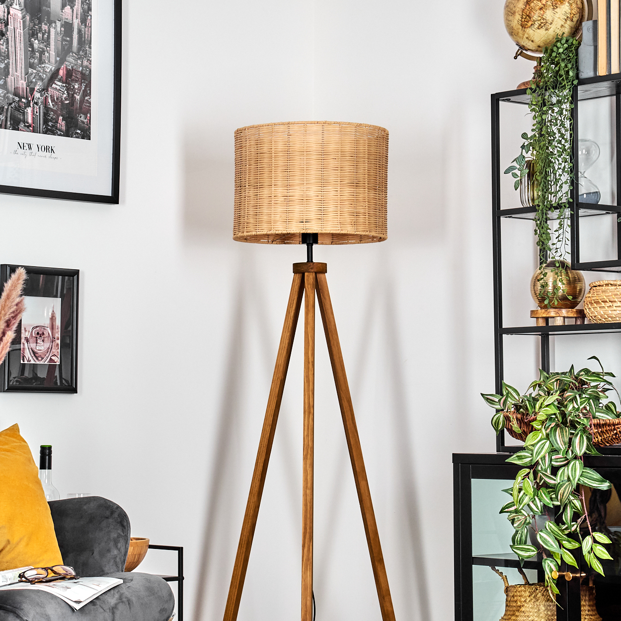 Ein gewebter Lampenschirm auf einem Holzstativ. Rechts ein schwarzes Regal mit Pflanzen und Deko. Links ein gerahmtes New York-Bild.