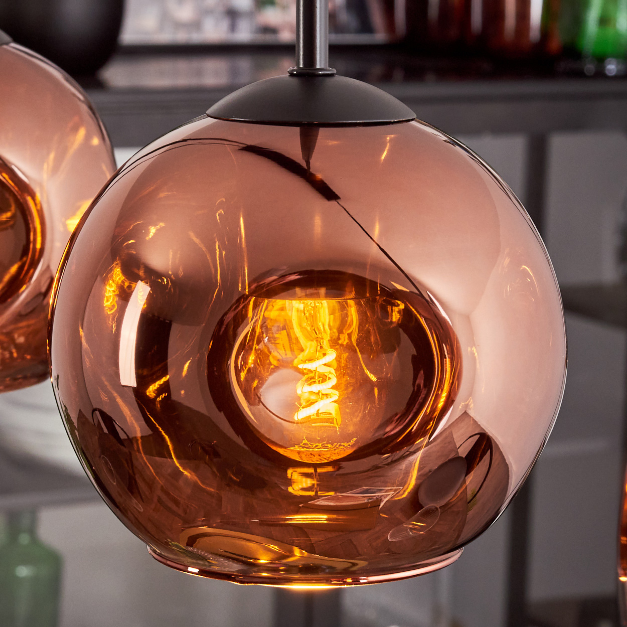 Nahaufnahme einer kupferfarbenen Glas-Pendelleuchte mit einer leuchtenden, spiralförmigen Glühbirne. Der Hintergrund ist unscharf.