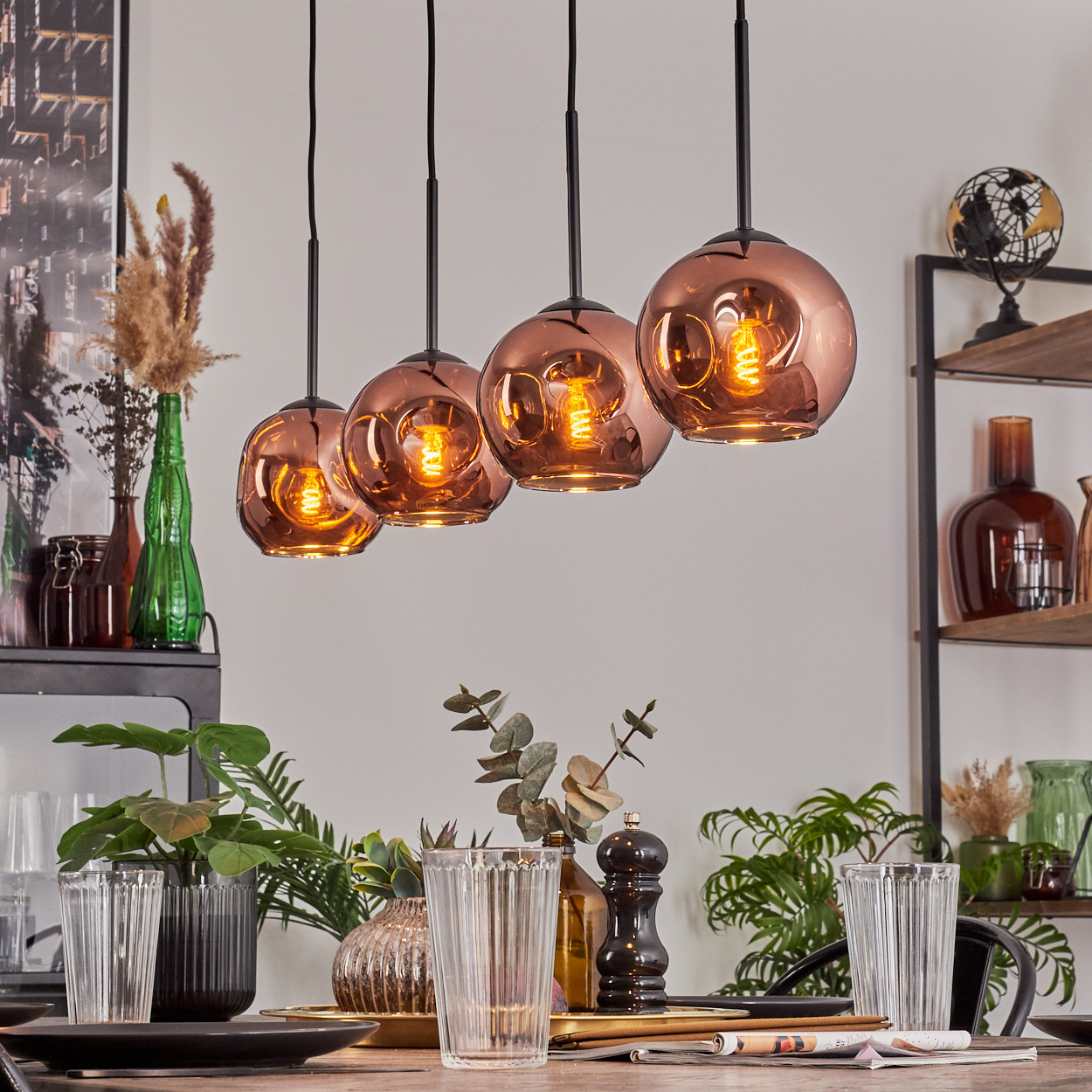 Vier kupferfarbene Kugelleuchten hängen über einem Tisch, der mit Gläsern und Pflanzen gedeckt ist. Regale mit Dekoration sind im Hintergrund.