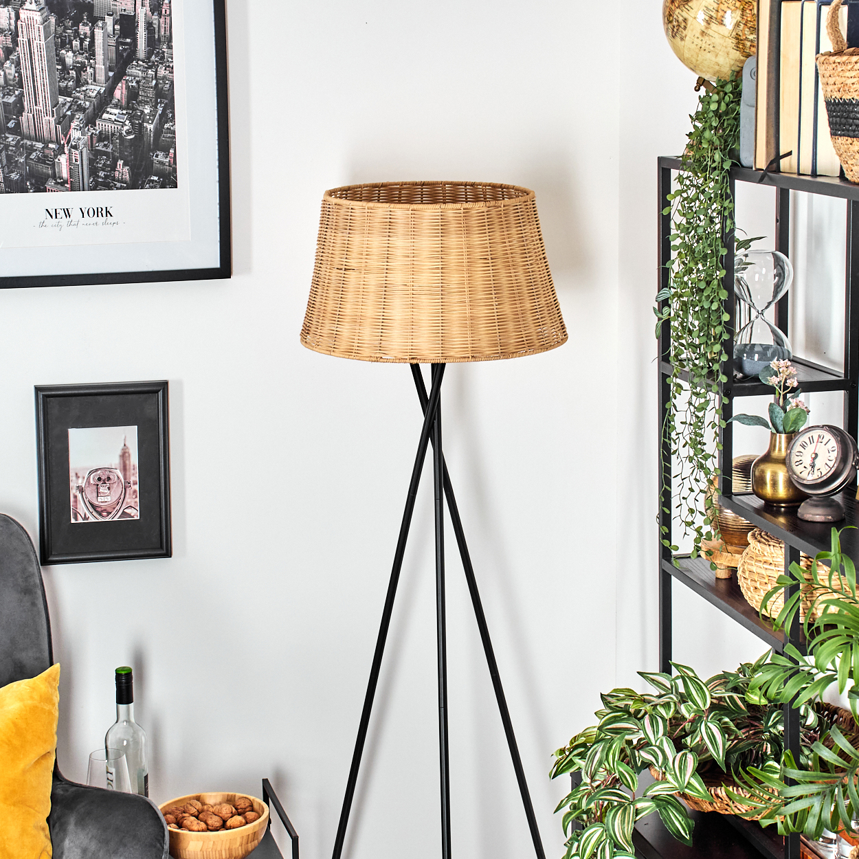 Eine gewebte Lampe mit schwarzen Beinen steht in einem Raum mit einem Regal voller Dekorationen und Pflanzen sowie gerahmten Bildern an der Wand.