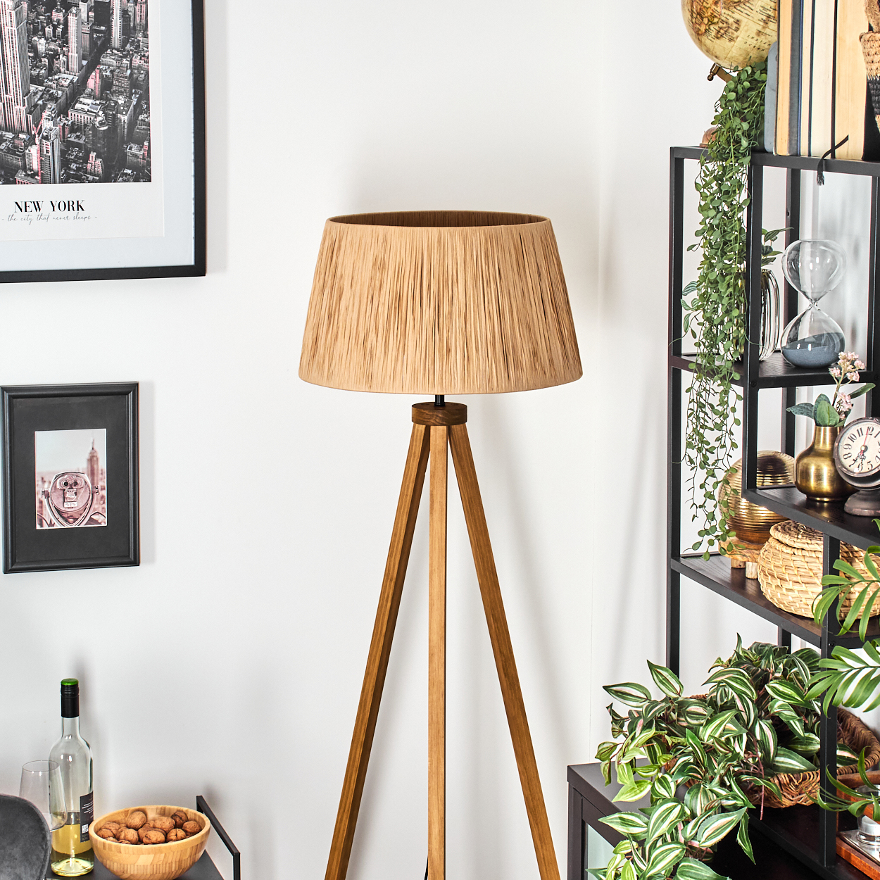 Ein Raum mit einer Holzlampe, schwarzen Regalen mit Dekoration, Pflanzen und gerahmten Bildern. Eine Schüssel mit Nüssen und eine Weinflasche stehen auf einem Tisch.