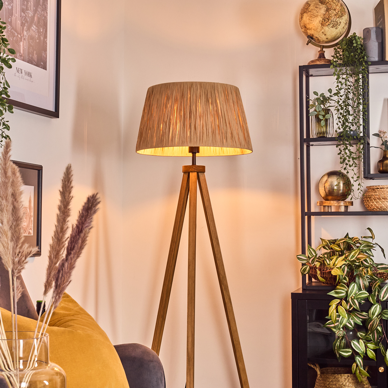 Eine hölzerne Stehlampe steht in einem Raum. Globus, Pflanzen und andere Dekorationen sind auf einem schwarzen Regal zu sehen. Ein gelbes Kissen ist zu sehen.