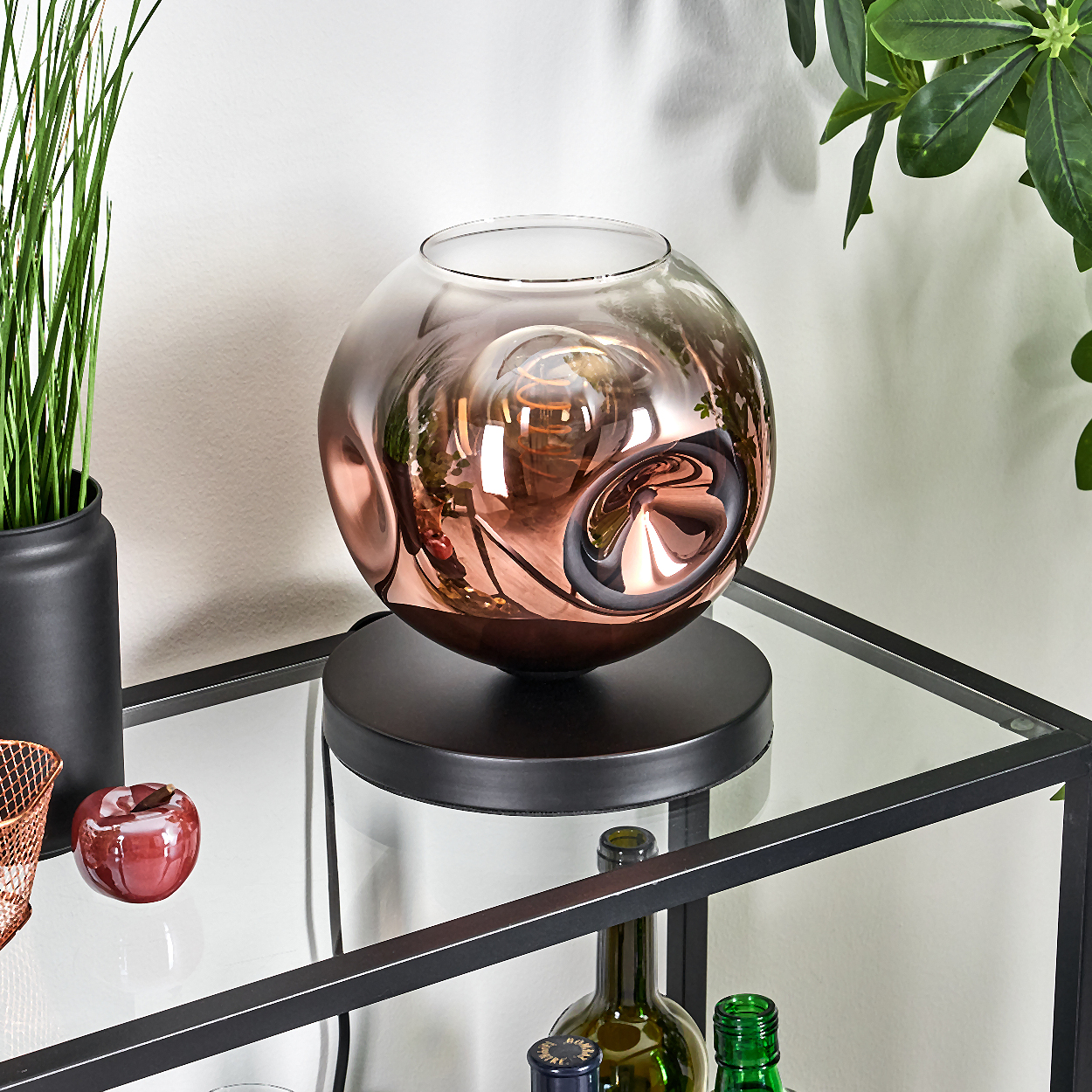 Eine dekorative Lampe mit einer kupferfarbenen Glaskugel steht auf einem Glasregal. Weitere Gegenstände sind Flaschen und ein roter Apfel.