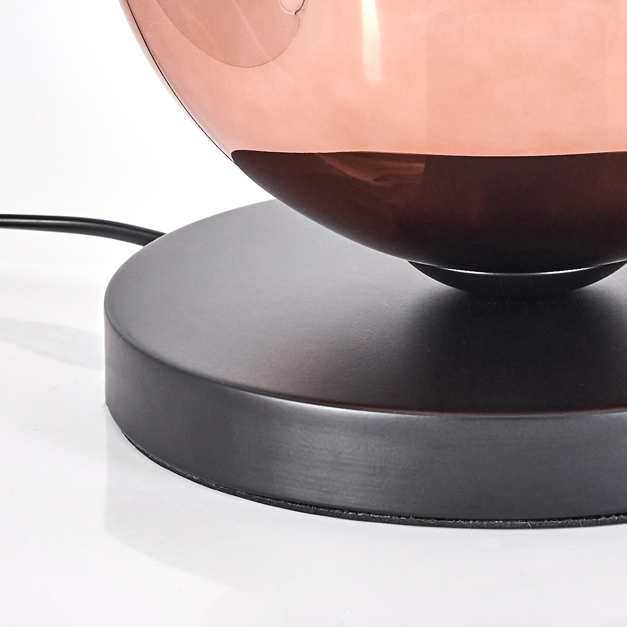 Eine moderne Lampe mit kupferfarbenem Schirm und schwarzem Sockel. Der Schirm reflektiert. Ein schwarzes Kabel ist sichtbar.